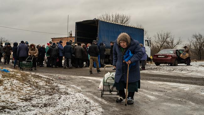 Auf einer Straße. Menschen in Winterkleidung stehen vor der offenen Ladefläche eines LKWs. Eine alte Frau mit Fellmütze und Gehstock kommt auf die Kamera zu, sie zieht eine kleine Transportrodel mit gefüllten Plastiksäcken. 