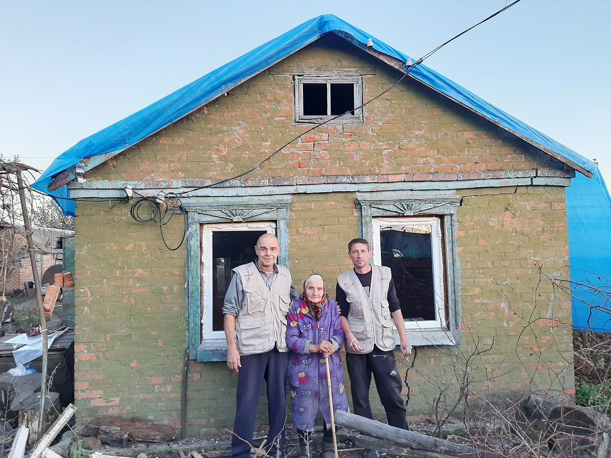 Eine alte Frau mit Gehstock steht vor einem brüchigen Haus mit blauer Plane über dem Dach. Zwei Männer stehen neben ihr, sie tragen Arbeitskleidung.