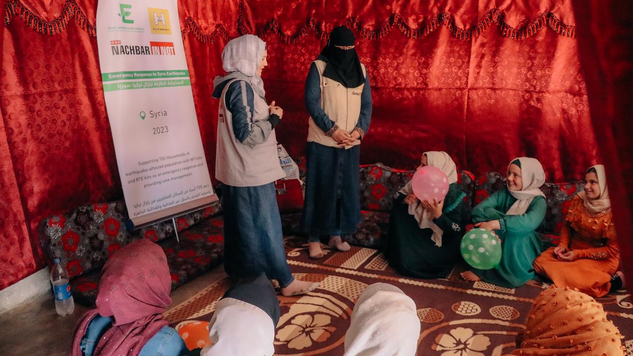 In einem Zelt mit roten Stoffwänden und Teppichen sitzt eine Gruppe von Mädchen auf dem Boden um zwei Frauen, die zu ihnen sprechen. Die Frauen und Kinder sind traditionell gekleidet und tragen Kopftücher. Die Stimmung scheint informell und aufmerksam zu sein.