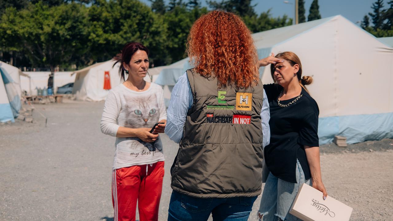 Drei Personen stehen auf einem Platz mit Zelten im Hintergrund und scheinen ein ernstes Gespräch zu führen. Eine der Frauen - sie hat rote lockige Haare - steht mit dem Rücken zur Kamera, auf ihrer Weste sind Logos von Hilfsorganisationen, darunter Hilfswerk und NACHBAR IN NOT, zu sehen.