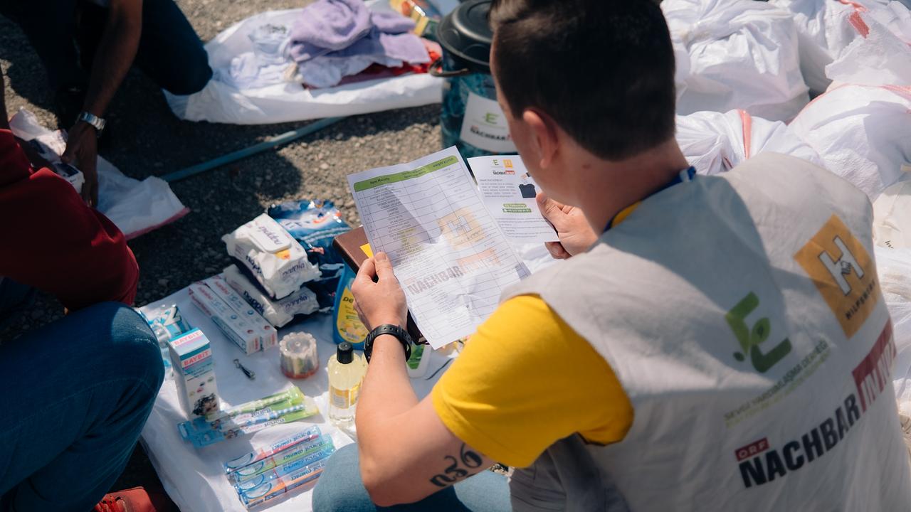 Eine Person in einer Weste mit Hilfswerk-Logo liest ein Dokument, umgeben von medizinischen und Hygieneartikeln, die auf einem weißen Tuch am Boden ausgelegt sind. Daneben sind gefüllte weiße Säcke vorbereitet.