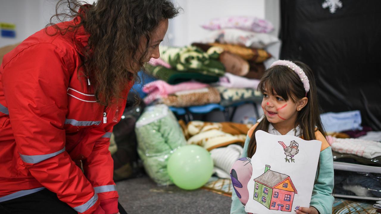 Eine Helferin in einer roten Jacke kniet vor einem kleinen Mädchen mit bunt bemaltem Gesicht. Das Mädchen hält eine Zeichnung von einem Haus und einem Vogel hoch. Sie scheinen eine herzliche Interaktion zu haben. Im Hintergrund sind Decken und Bettwäsche zu erkennen, was auf eine Notunterkunft hinweist.