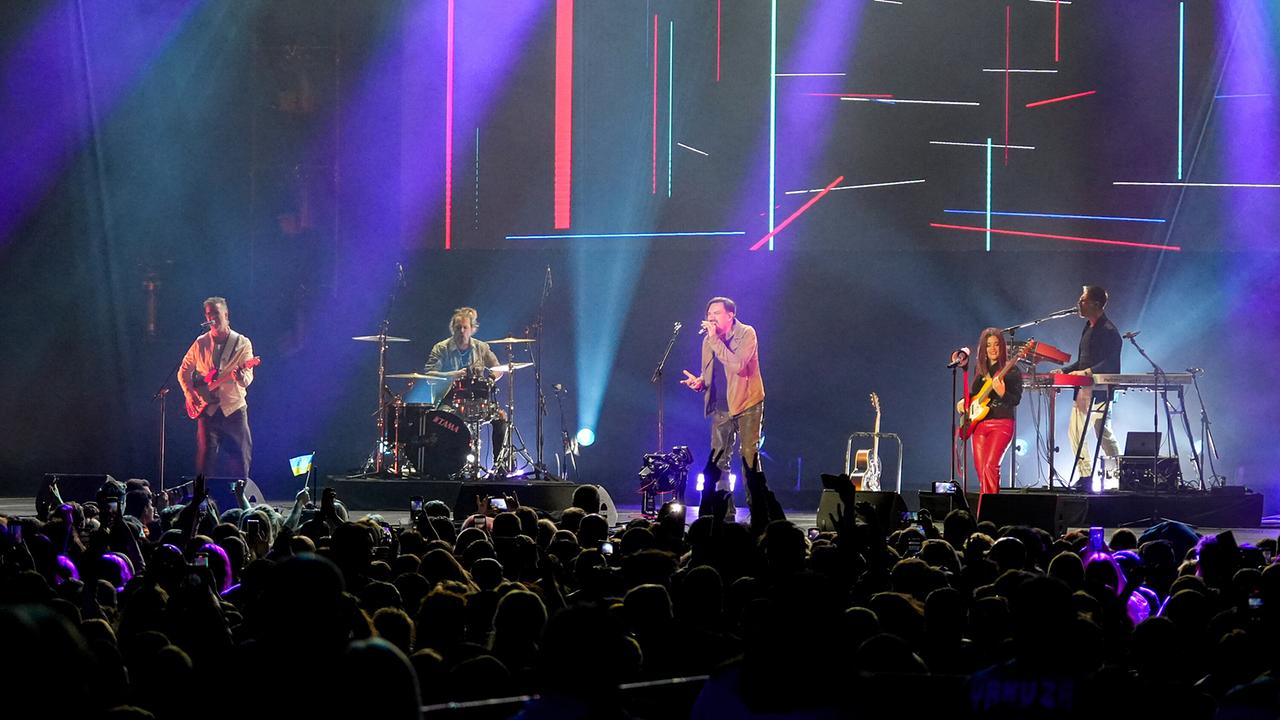 Ein Gitarrist, ein Schlagzeuger, ein Sänger, ein Bassist und ein Keyboard-Spieler auf der Bühne vor einer Menschenmenge.