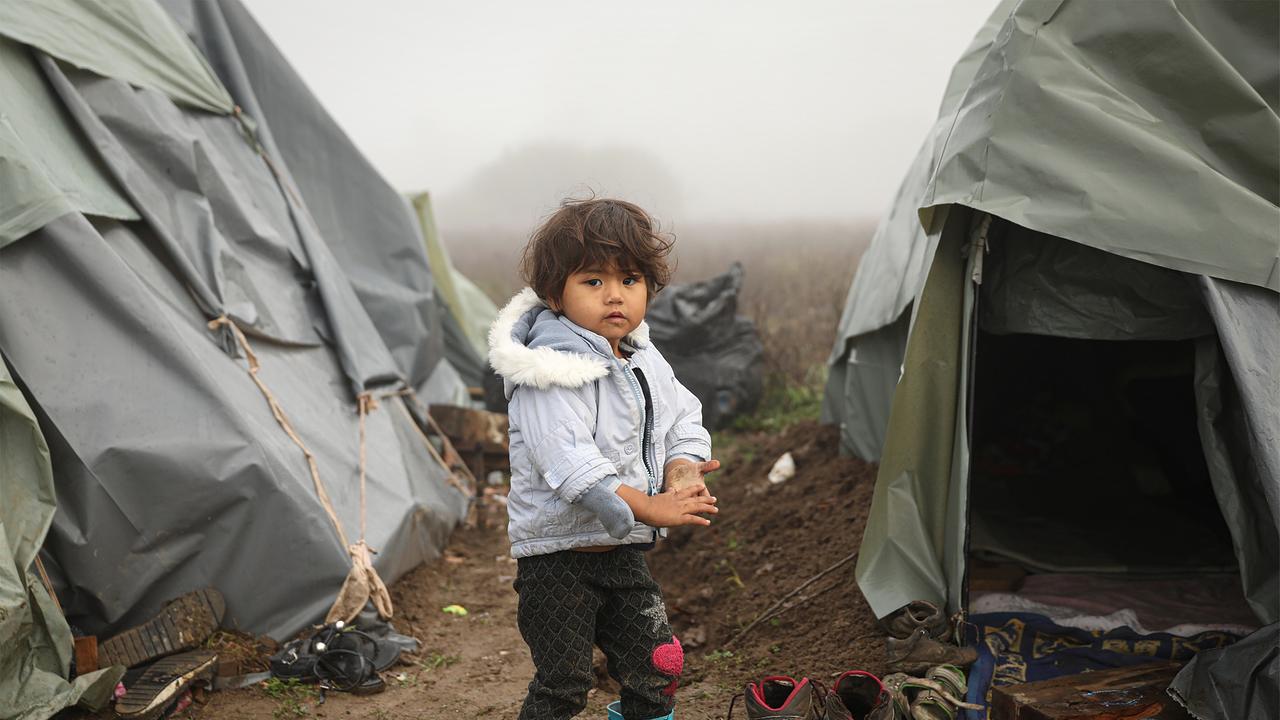 Ein Kleinkind steht in Gummistiefeln und Winterjacke im Schlamm zwischen Zelten. Man sieht Müllsäcke und dreckige, nasse Schuhe. Teilweise sind es offene Sommerschuhe.