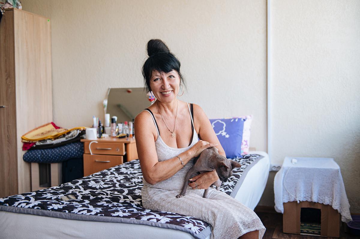 Eine Frau, Mitte 50, mit dunklen Haaren in einem Dutt, sitzt auf einem Bett und lächelt. Auf ihrem Schoß sitzt eine graue Nacktkatze. Auf dem Nachttisch im Hintergrund stehen Hygieneartikel, daneben liegen mehrere zusammengelegte Decken.