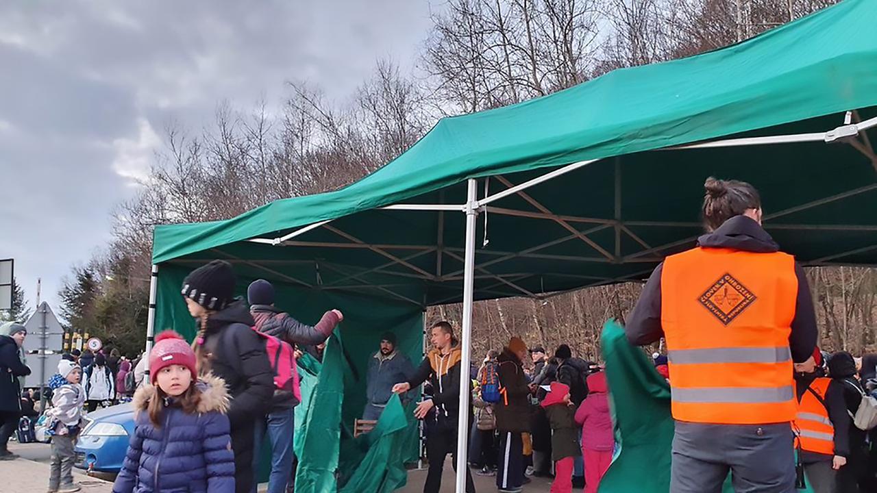 Mitarbeiter einer Hilfsorganisation bauen ein großes Zelt auf. Unter dem Zelt stehen Familien mit Kindern.