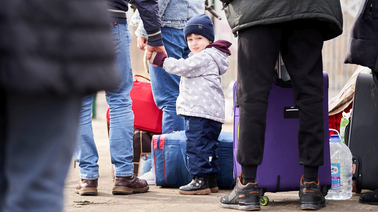 Man sieht Menschen von der Hüfte abwärts neben ihren Koffern stehen. In der Mitte des Bildes hält ein Kind die Hand eines Mannes, es schaut in die Kamera.