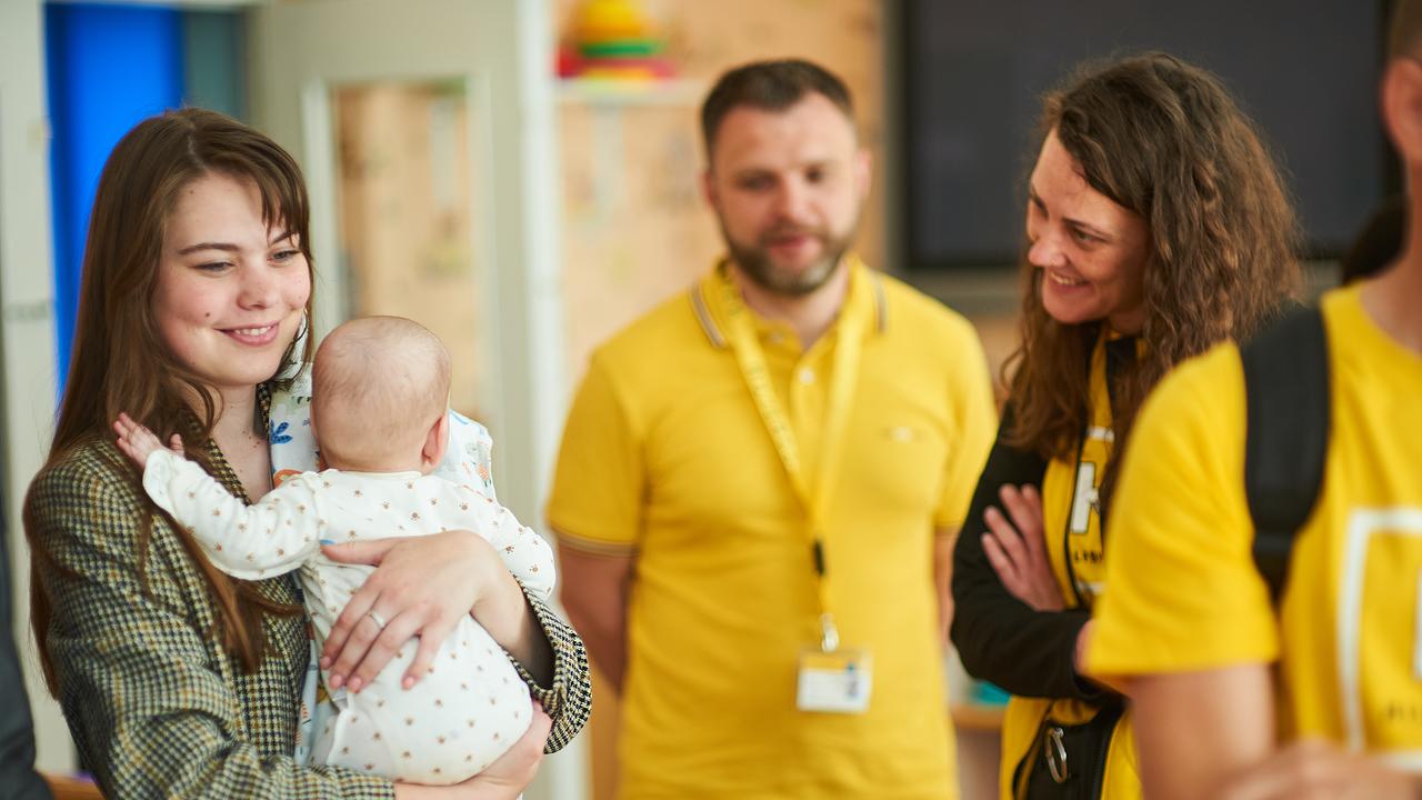Drei Menschen sind auf dem Bild abgebildet. Von der Organisation "Hilfswerk" sind zwei Mitarbeiter:innen, eine Frau und ein Mann, in gelbe T-Shirts auf der rechten Bildseite zu sehen. Die Mitarbeiterin lächelt eine Frau auf der linken Bildseite an. Die Frau hat braune Haaren und hält ein Baby in der Hand. Sie schaut Richtung Boden und lächelt. 