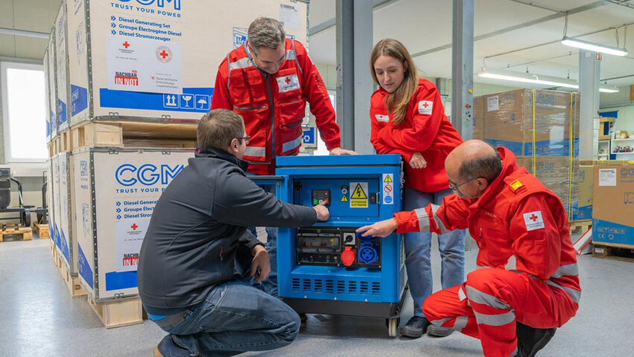 Mitglieder des Österreichischen Roten Kreuzes sehen sich in einer Lagerhalle einen blauen Generator an.