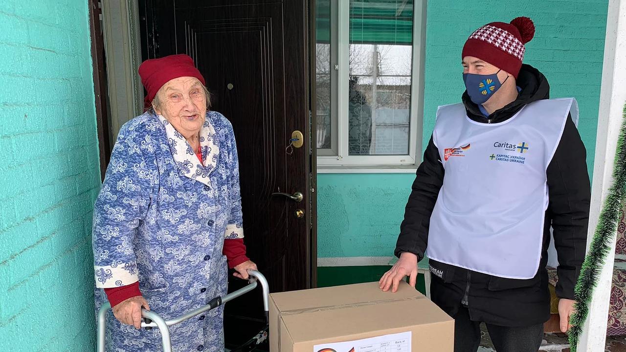 Auf dem Bild sind eine ältere Dame mit Gehhilfe sowie ein Helfer zu sehen. Der Mann bringt der Frau zwei große Kisten. 