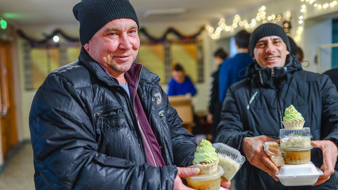 Zwei Männer in Winterkleidung stehen in einem Innenraum. Beide halten einen kleinen Turm warmes Essen in Einweggeschirr in der Hand, darauf ein Cupcake. Die beiden Männer lächeln.