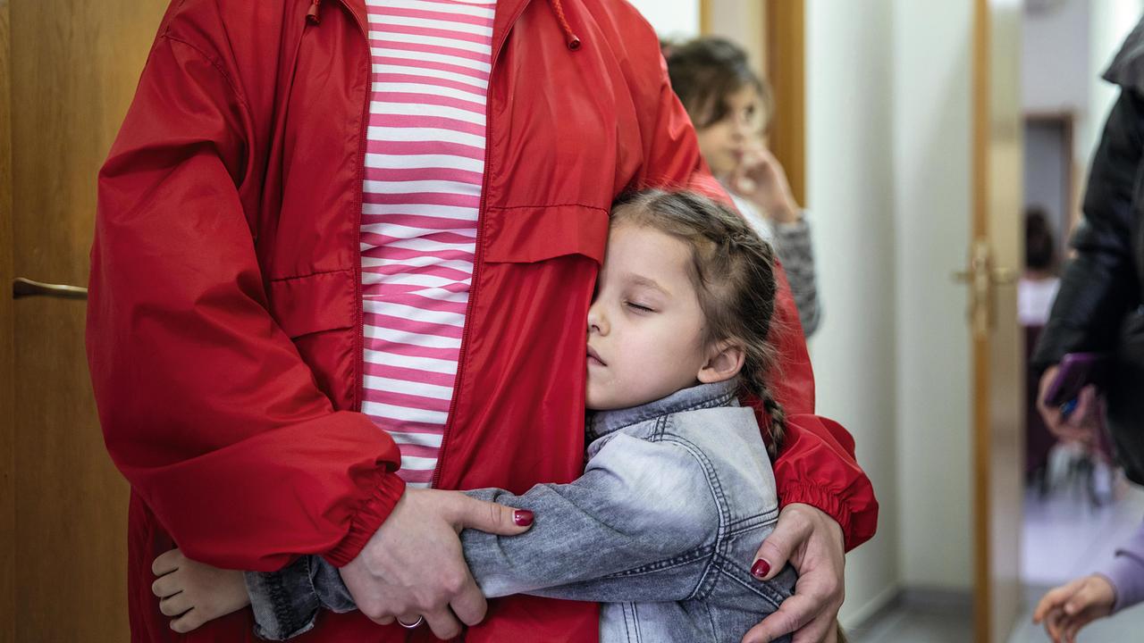 Ein junges Mädchen mit Zöpfen umarmt eine Frau in einer roten Caritas-Jacke. Ihre Augen sind geschlossen. Die Frau sieht man nur von den Schultern abwärts, das Mädchen geht ihr bis zur Hüfte.