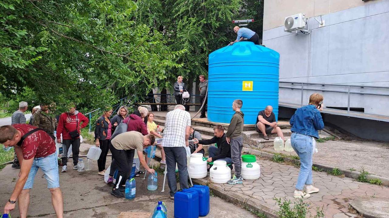 Auf einer Straße stehen rund 15 Menschen mit großen Behältern vor einem mehrere Meter hohen Wassertank. Der Wassertank ist blau mit einem gelben Aufkleber. Der Aufkleber ist das Hilfswerk-Logo.