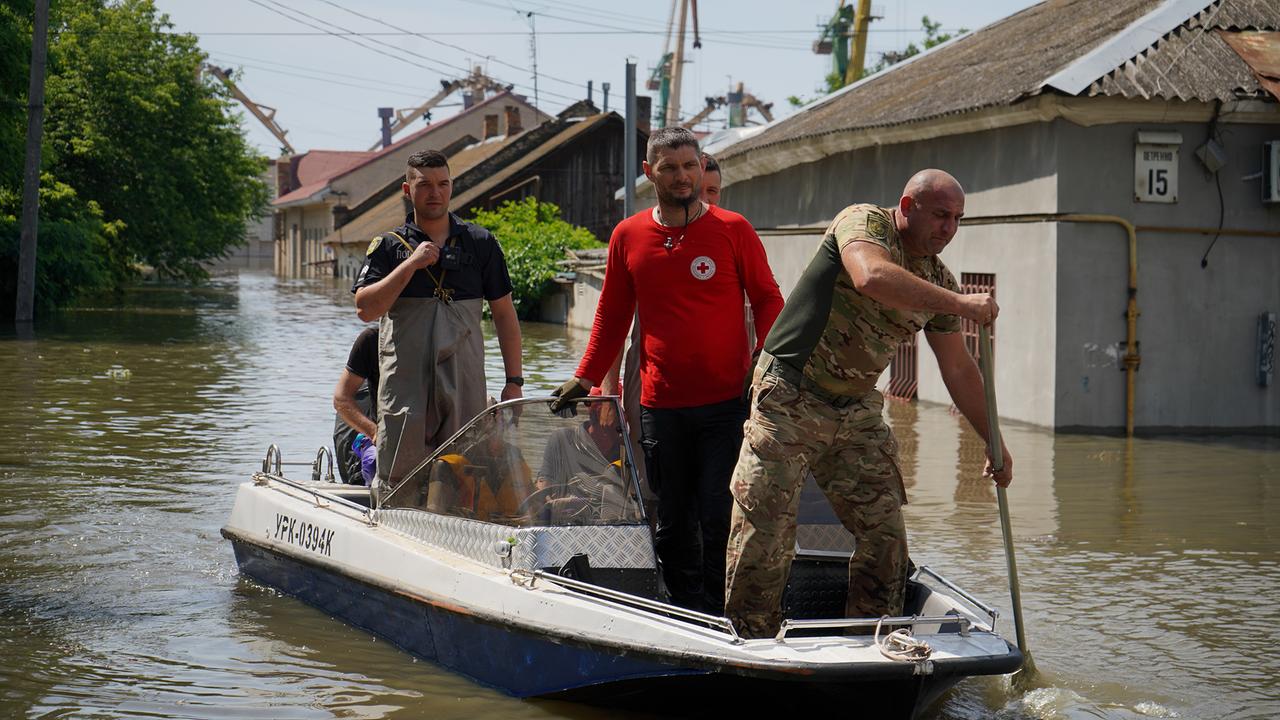 Eine überschwemmte Straße. Mehrere Männer, darunter Rotkreuz-Mitarbeiter und Soldaten, sind in einem kleinen Elektroboot unterwegs.
