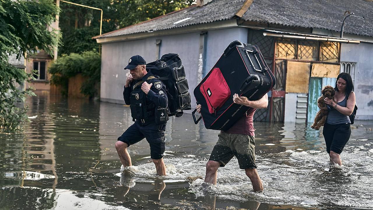 Zwei Männer tragen große Koffer über das Wasser. Das Wasser reicht bis zu den Schienbeinen. Die Häuser im Hintergrund stehen unter Wasser. 