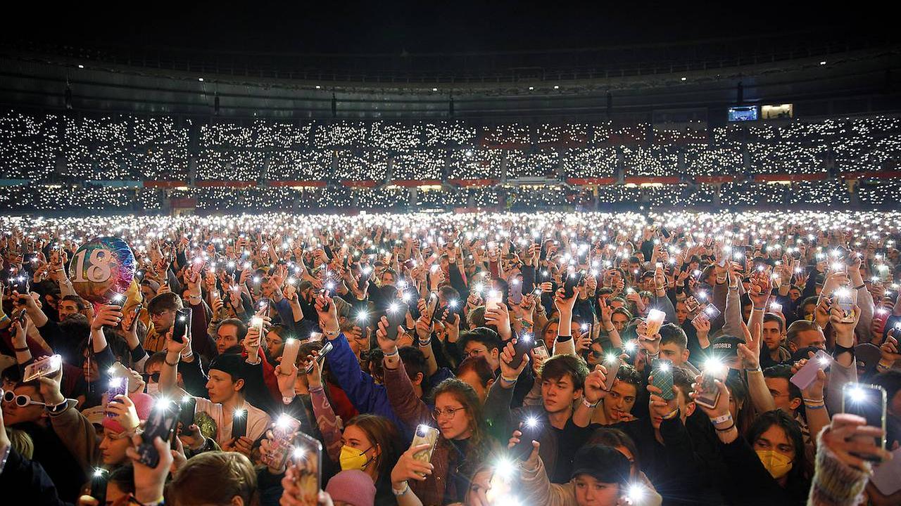 Eine Menschenmasse steht vor der Bühne, im Dunklen sind zigtausende Lichter von Handytaschenlampen zu sehen. 