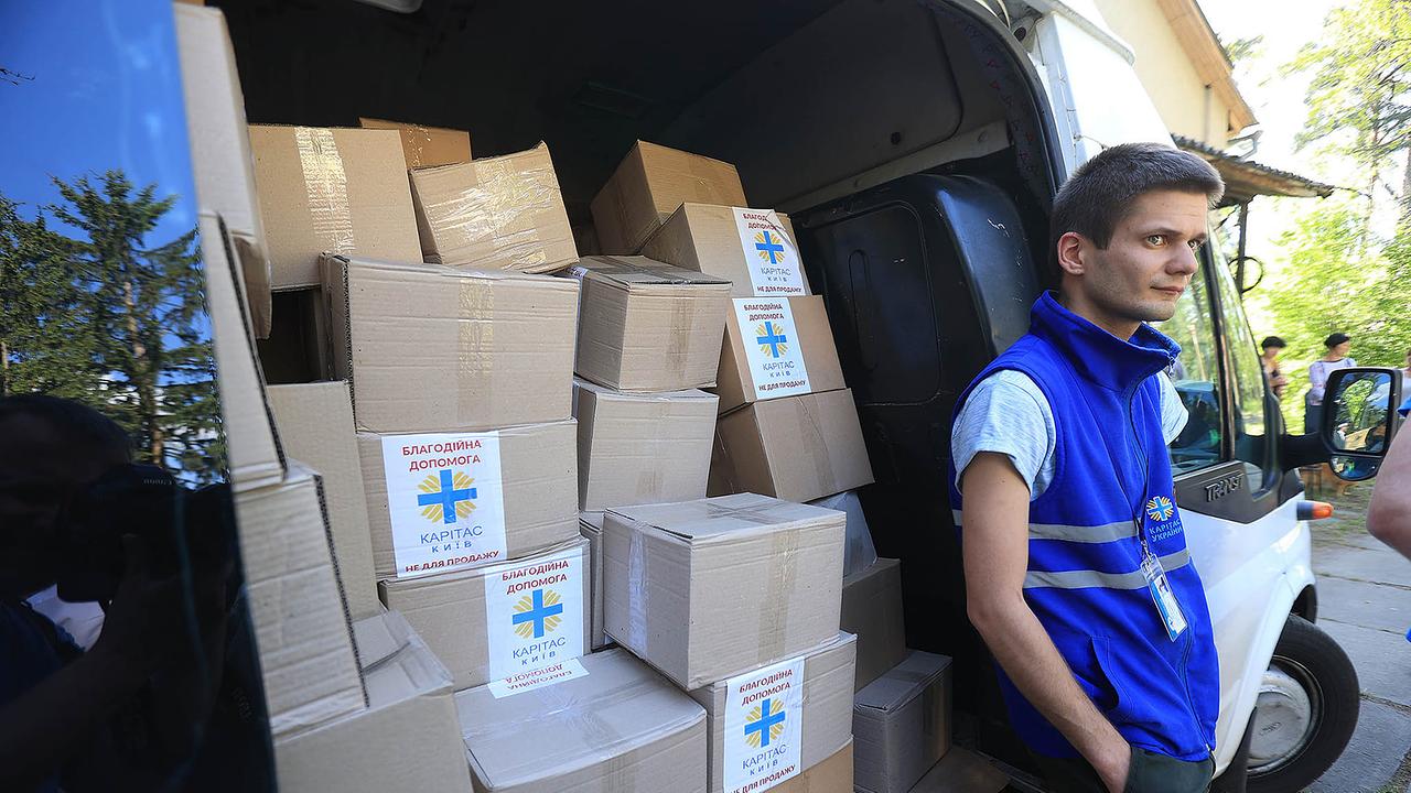 Ein junger Helfer der Caritas in einer blauen Weste steht vor einem weißen Lieferwagen. Die Schiebetür ist offen, darin befinden sich dutzende Hilfspakete.