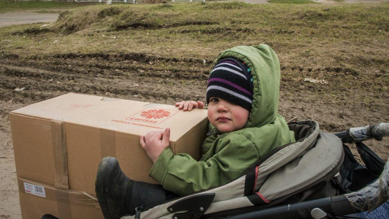 Ein kleines Kind sitzt mit grünem Kapuzenpullover, einer gestreiften Mütze und dicken Winterstiefeln in einem Kinderwagen und hält ein Caritas-Hilfspaket.