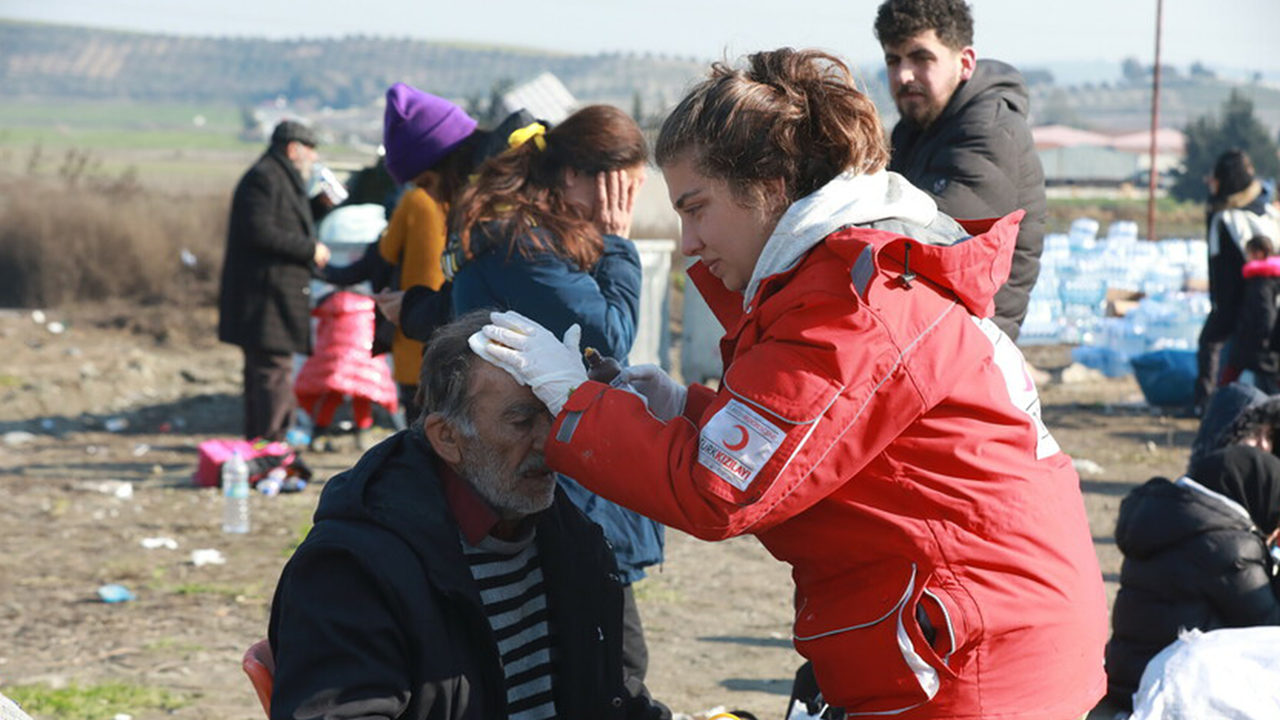 Auf dem Bild verarztet eine junge Frau mit roter Jacke einen älteren Mann. Die Frau trägt weiße Handschuhe und hält eine Hand auf den Kopf des Mannes. Im Hintergrund sind weitere Menschen im Freien zu sehen. 