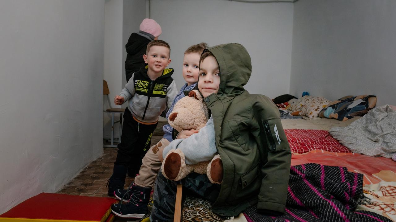 Drei junge Kinder sitzen in einem provisorischen Wohnraum auf einem Bett. Die Kinder tragen Winterkleidung, einer hält ebenfalls einen Teddybären. Sie blicken neugierig in die Kamera, umgeben von Bettwäsche und persönlichen Gegenständen, was auf eine temporäre Unterbringung hinweist.