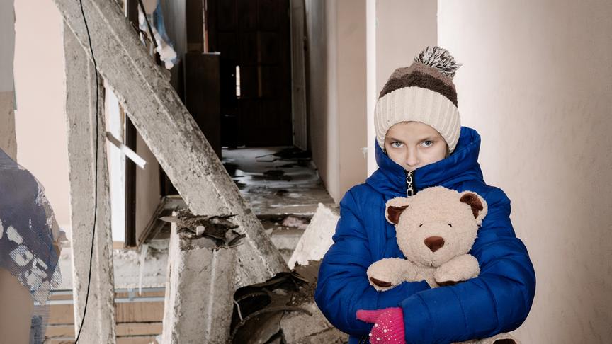 Ein Kind in einem blauen Mantel und einer Mütze hält einen Teddybären fest und steht vor den Trümmern eines zerstörten Gebäudes. Es blickt direkt in die Kamera, sein Gesichtsausdruck ist ernst.