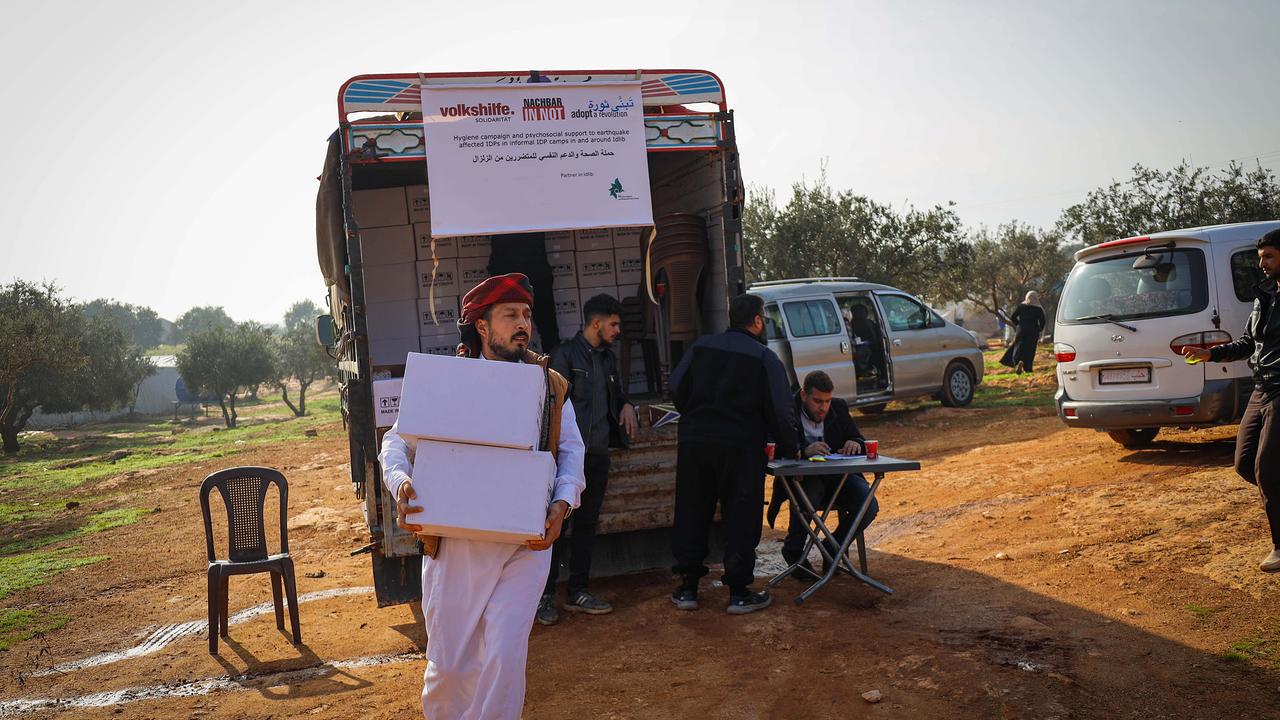Ein Mann mit einem roten Kopftuch trägt mehrere weiße Kisten aus einem Lastwagen, vor dem ein weißes Banner und ein Tisch stehen, an dem zwei Männer sitzen. Sie befinden sich in einem ländlichen Gebiet mit Bäumen und einigen Fahrzeugen im Hintergrund. Die Szene zeigt eine geordnete Verteilung von Hilfsgütern mit einem Gefühl der Kooperation.