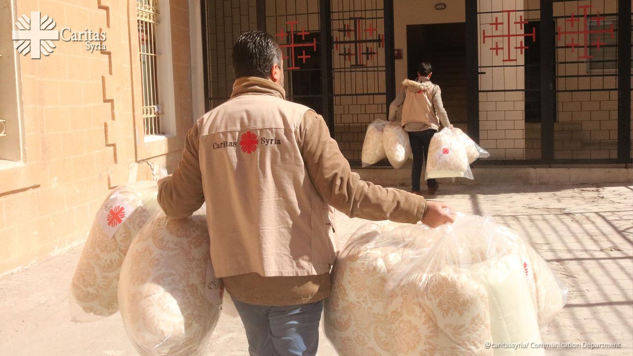 Zwei Männer tragen große, verpackte Gegenstände, die wie Bettdecken aussehen, an einem sonnigen Tag vor einem Gebäude mit Kreuz-Symbolen an den Fenstern. Ihre Jacken tragen das Logo der Caritas Syria. Es herrscht eine Atmosphäre des geschäftigen Helfens.