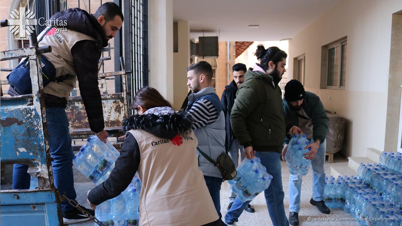 Mehrere Personen laden Wasserflaschen aus einem Lieferwagen in einem Innenhof ab. Sie tragen Westen mit dem Logo der Caritas Syria und sind dabei, Wasserflaschen an der Wand aufzustapeln.