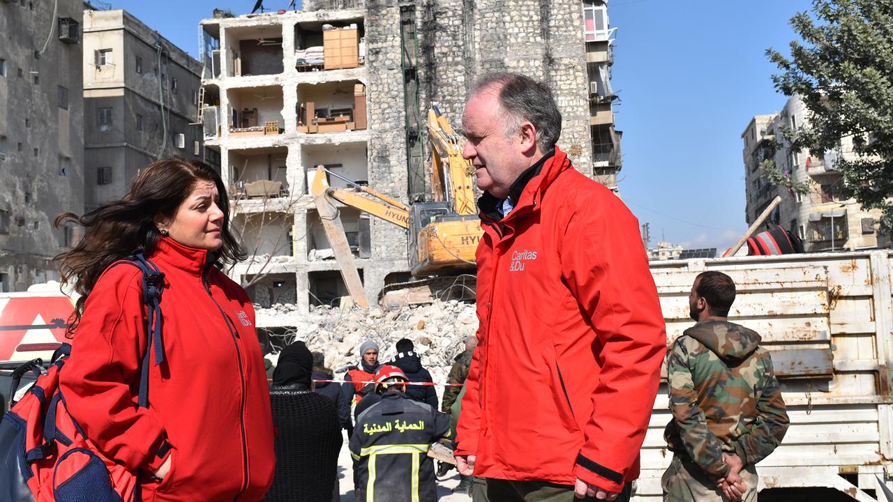 Eine Frau und ein Mann (Andreas Knapp) in roter Kleidung mit Logos de Caritas stehen in einer zerstörten städtischen Szenerie und führen ein Gespräch. Hinter ihnen sind ein Bagger und zerstörte Gebäude zu sehen,