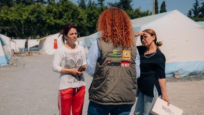 Drei Personen stehen auf einem Platz mit Zelten im Hintergrund und scheinen ein ernstes Gespräch zu führen. Eine der Frauen - sie hat rote lockige Haare - steht mit dem Rücken zur Kamera, auf ihrer Weste sind Logos von Hilfsorganisationen, darunter Hilfswerk und NACHBAR IN NOT, zu sehen.