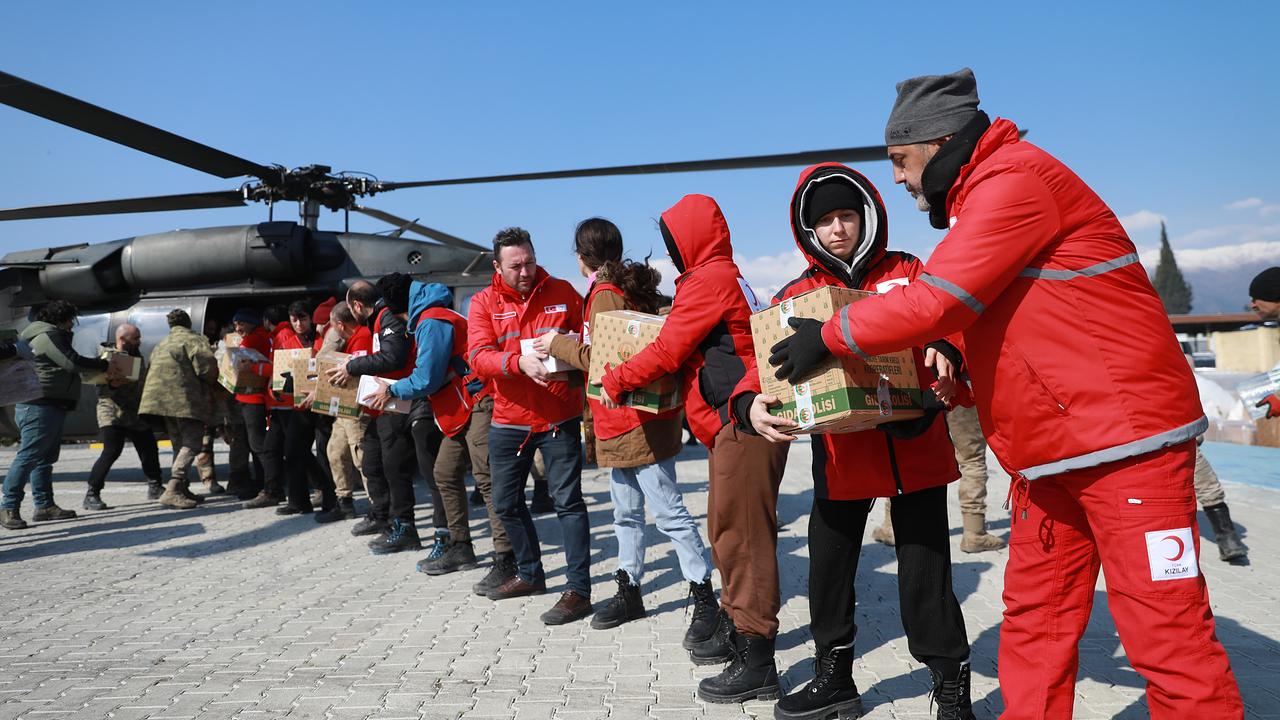 Mehrere Personen in roten Jacken stehen in einer Reihe und übergeben neben einem Hubschrauber Kisten aneinander. Sie stehen auf einem gepflasterten Platz, die Szene wirkt geordnet und geschäftig. Die Personen sind warm gekleidet.