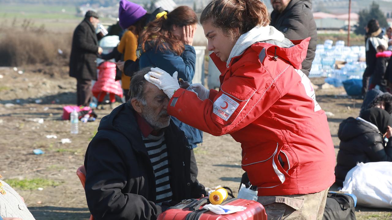 Eine Helferin in einer roten Jacke versorgt einen älteren Mann medizinisch, indem sie eine Behandlung an seinem Kopf vornimmt. Sie sind im Freien, und im Hintergrund sind andere Menschen und Hilfsgüter zu sehen.