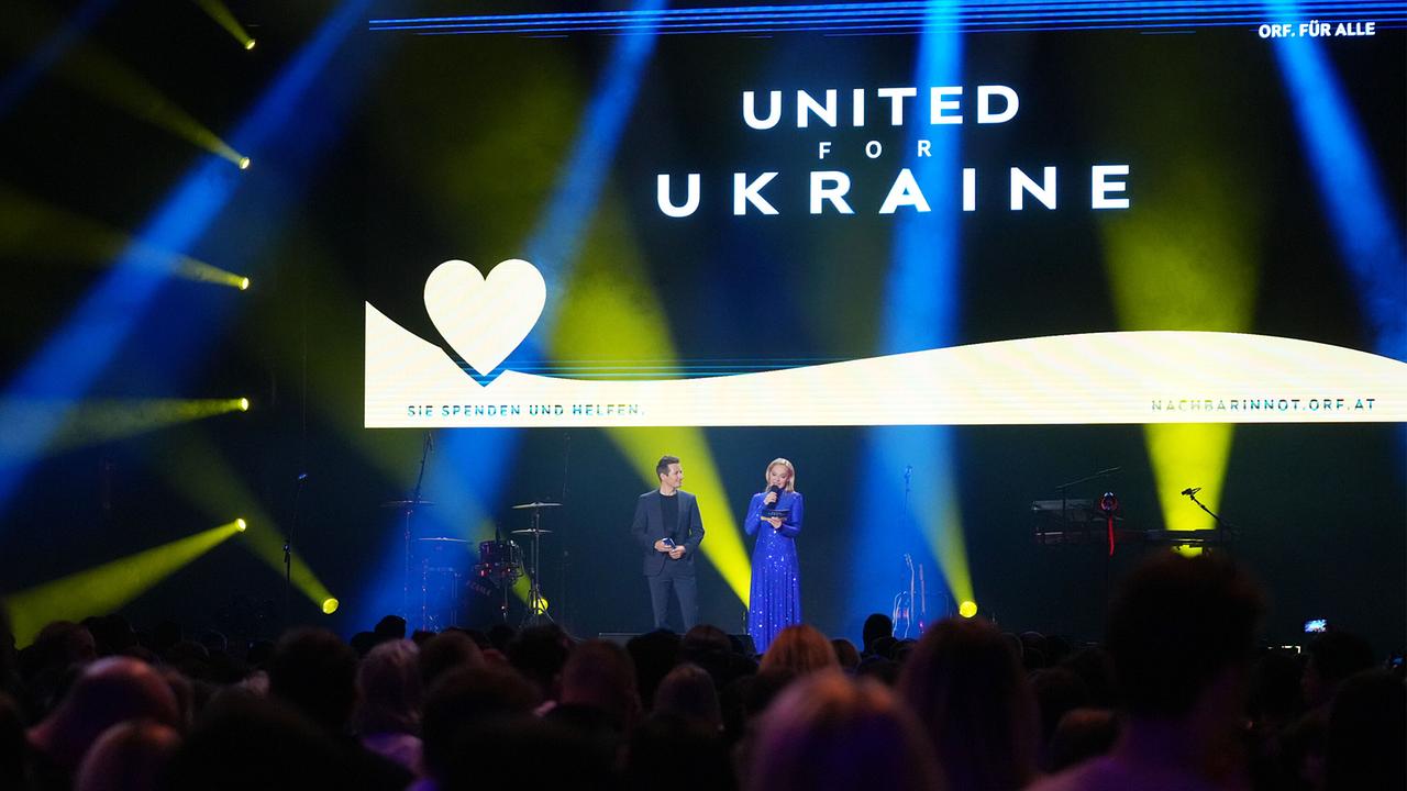 Das Foto zeigt zwei Personen, eine Frau und einen Mann, die auf einer Bühne stehen und zu einem Publikum sprechen. Der Mann trägt einen dunklen Anzug und kurze dunkle Haare, die Frau ist blond und trägt ein glitzerndes blaues Kleid. Sie sind vor einem großen Bildschirm positioniert, der die Worte "UNITED FOR UKRAINE" und ein Herzsymbol zusammen mit einem Aufruf zum Spenden zeigt. Die Bühne ist in dramatisches blaues und gelbes Licht getaucht, die Farben der ukrainischen Flagge, und das Publikum im Vordergrund ist verschwommen.