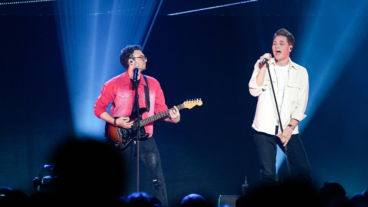 Zwei junge Männer stehen auf einer Bühne, beide haben ein Mikrofon vor sich. Der linke trägt ein rotes Hemd und spielt auf einer Gitarre, der rechte trägt ein offenes weißes Hemd über einem T-Shirt und singt. Beide haben helle Haut und kurze dunkle Haare.